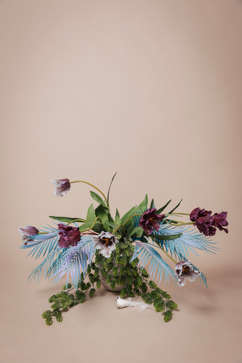 Boeket van luxe kunstbloemen met paarse tulpen en witte anjers, verrijkt met tropische blauwe palmbladeren en weelderig groen.