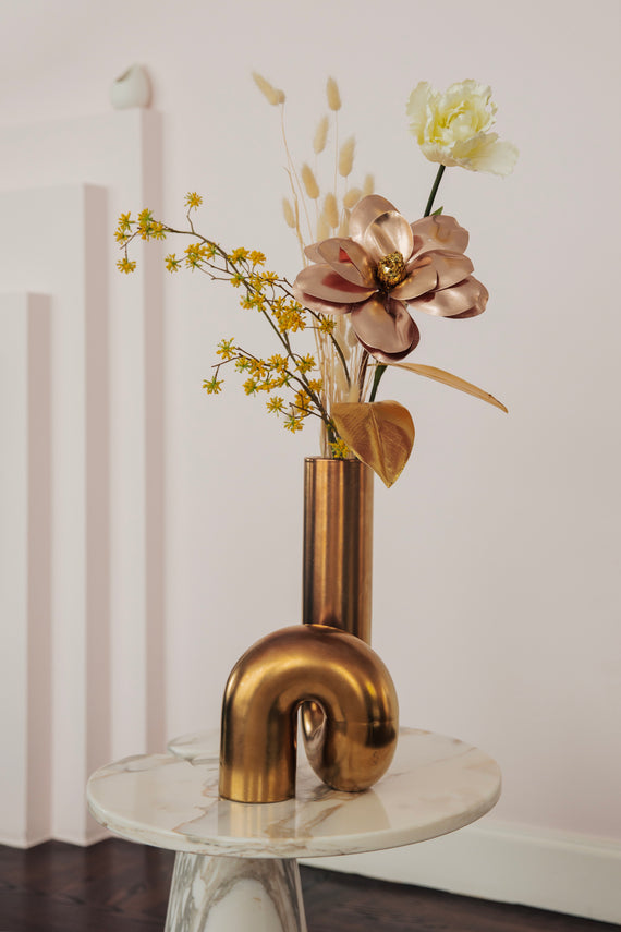 Een stijlvol kunstbloemarrangement in een gouden vaas op een marmeren tafel, met een gouden bloem, witte ranonkel en subtiele gele accenten.