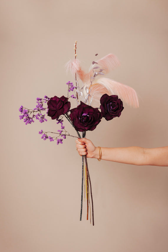 Een klein kunstboeket met dieprode rozen, paarse accentbloemen en zachte roze veren, samengebracht in een elegante samenstelling